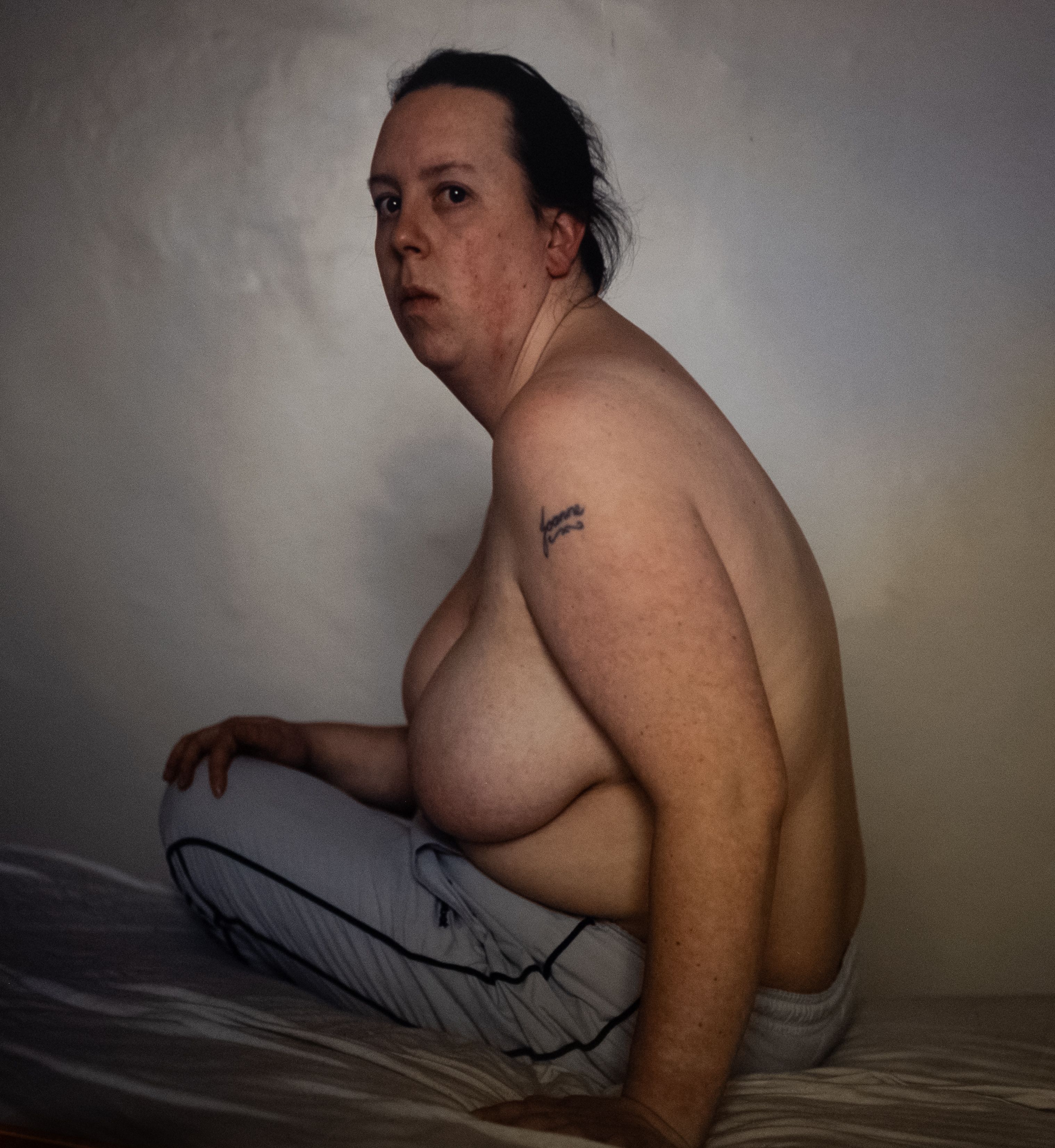 Joanne – A photograph of a topless women sitting on a bed, side profile. She has a tattoo on her upper arm reading ‘Joanne’ and wears white tracksuit trousers.