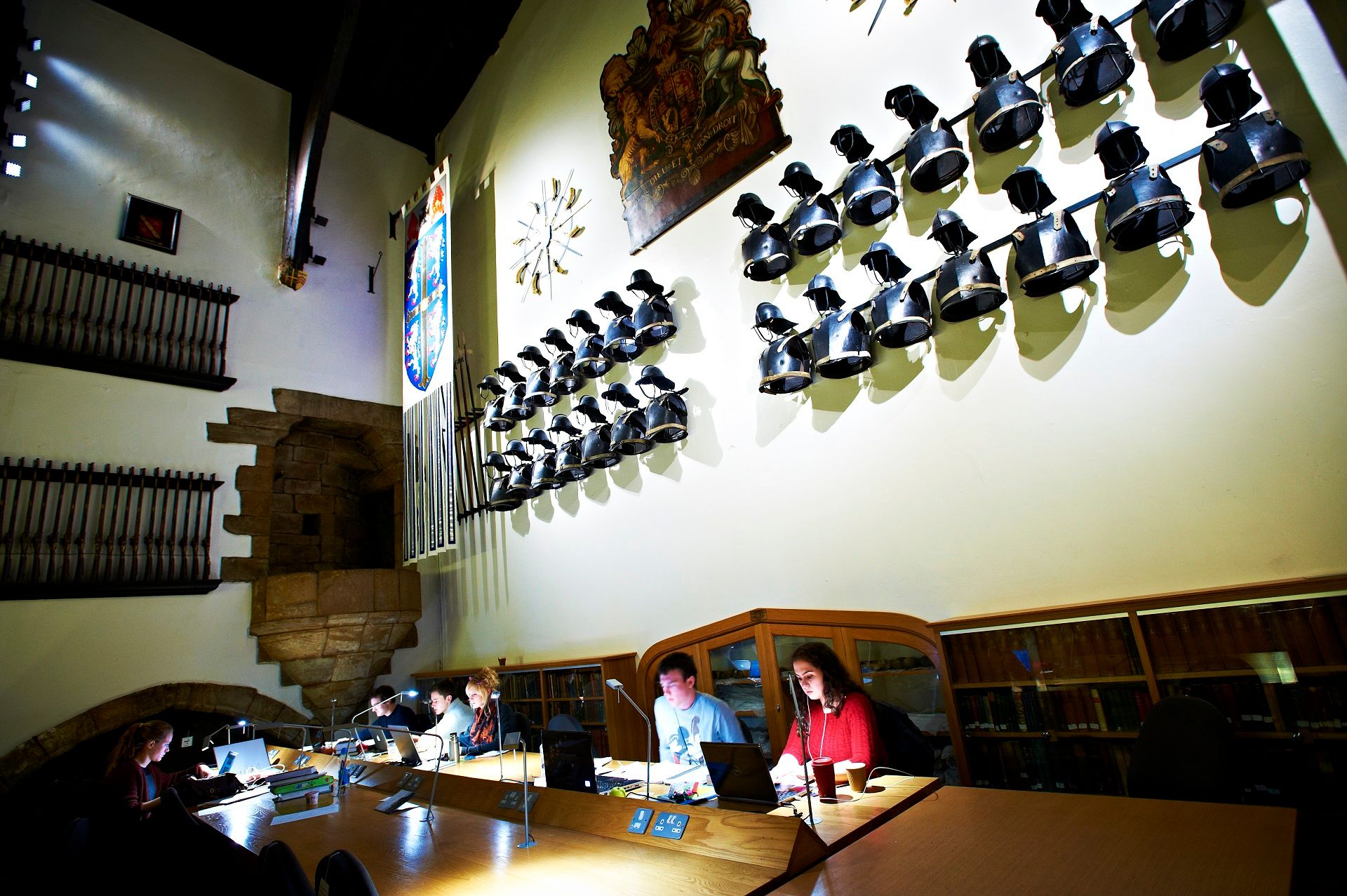 A coloured photograph taken in the Minstrel’s Gallery, looking towards the south wall of the Great Hall. At the top of the image are the armour. Below students are studying.