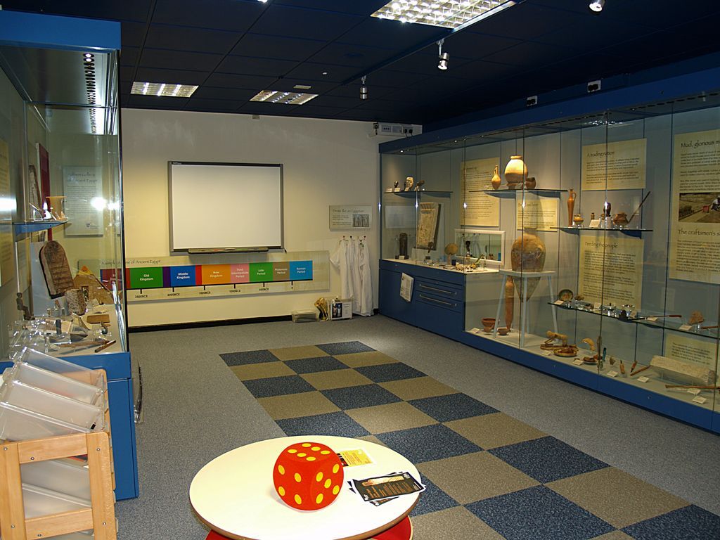 View of displays of ancient Egyptian objects around the edges of the Wolfson Gallery of ancient Egypt with white board on end wall, dressing up and game board marked out in tiles on the floor