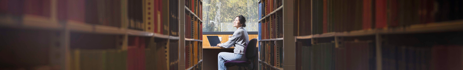 A young woman at the end of a long passage of library books sits at a library window looking up.