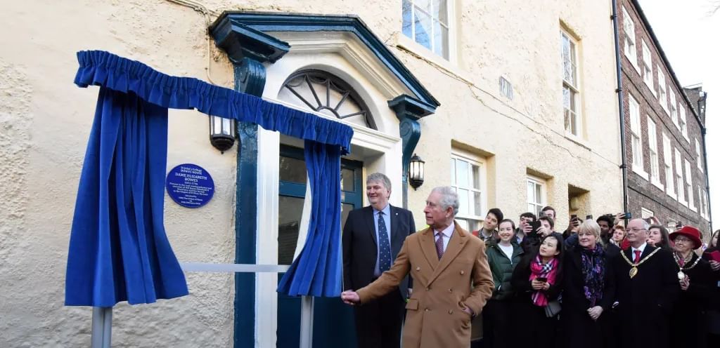 Man in brown coat pulls back curtain to reveal blue plaque on wall