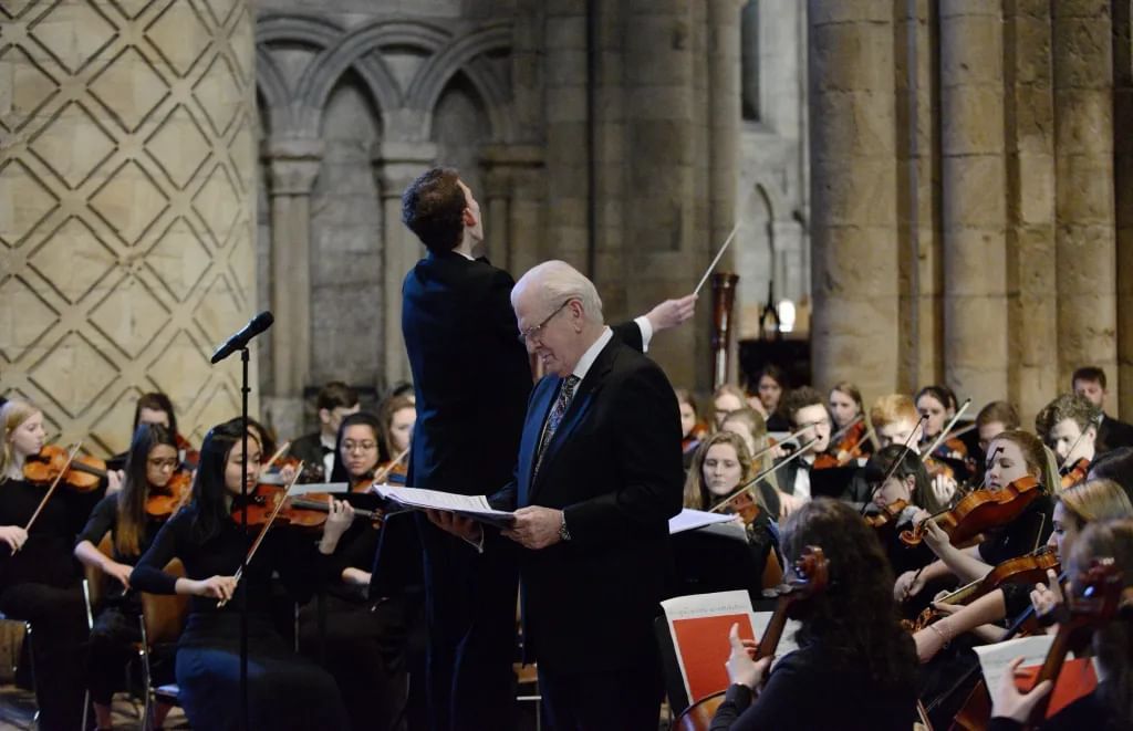 A white haired man looks down at a score, while a conductor behind him leads an orchestra