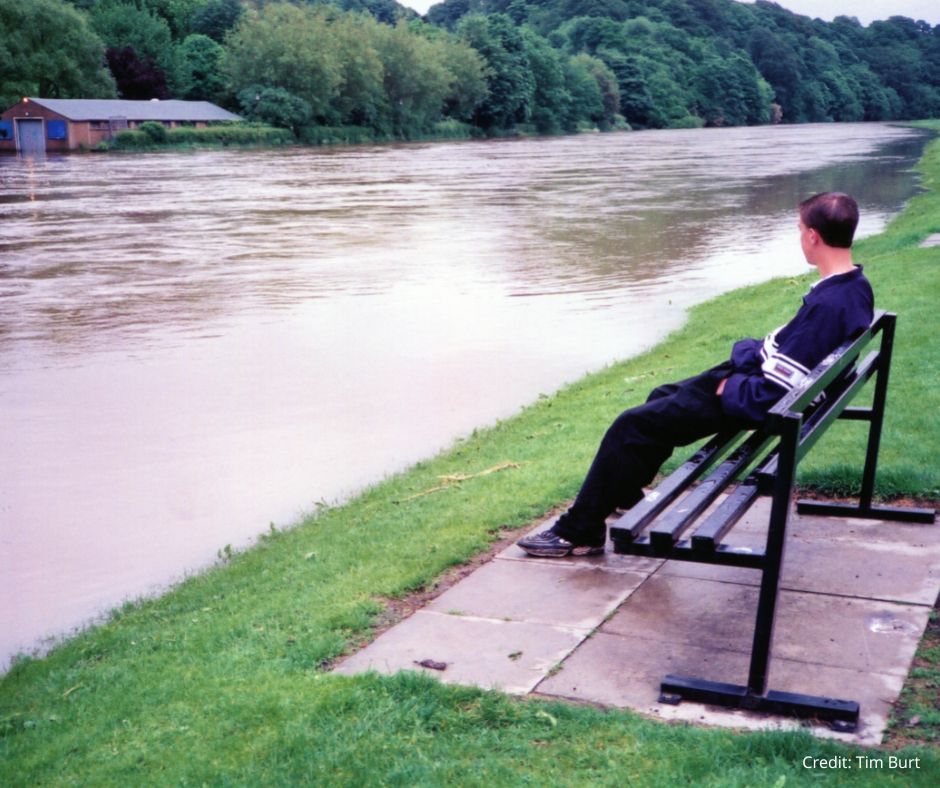 A student sits on a bench next to the flooded River Wear