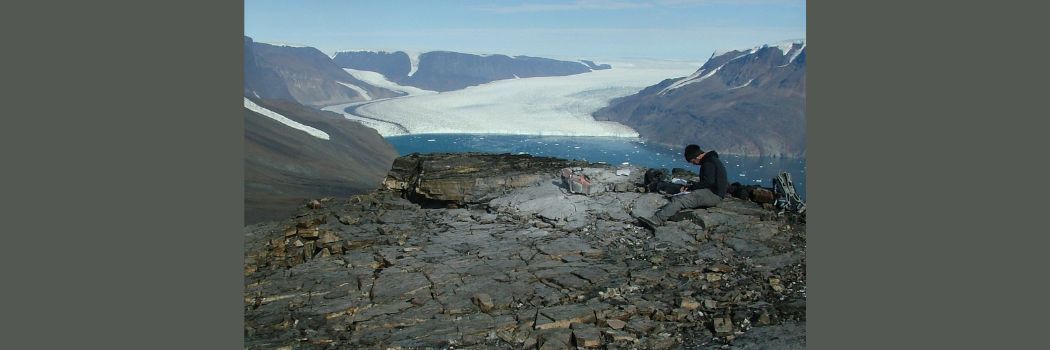 A scientist sits on rocks in the foreground with a huge white ice sheet in the background.
