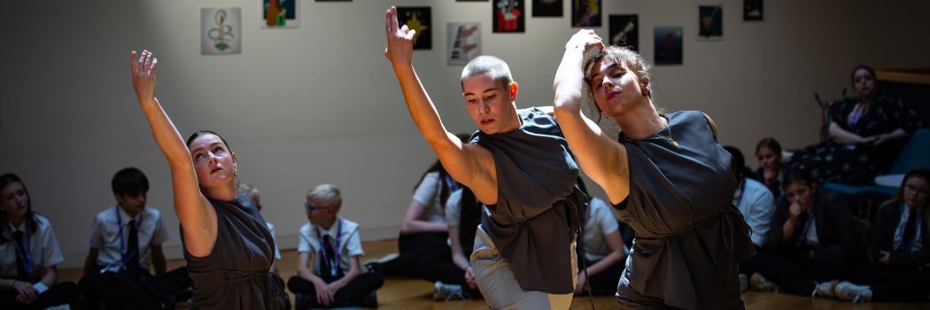 Three dancers perform as school children sit around them on the floor, watching