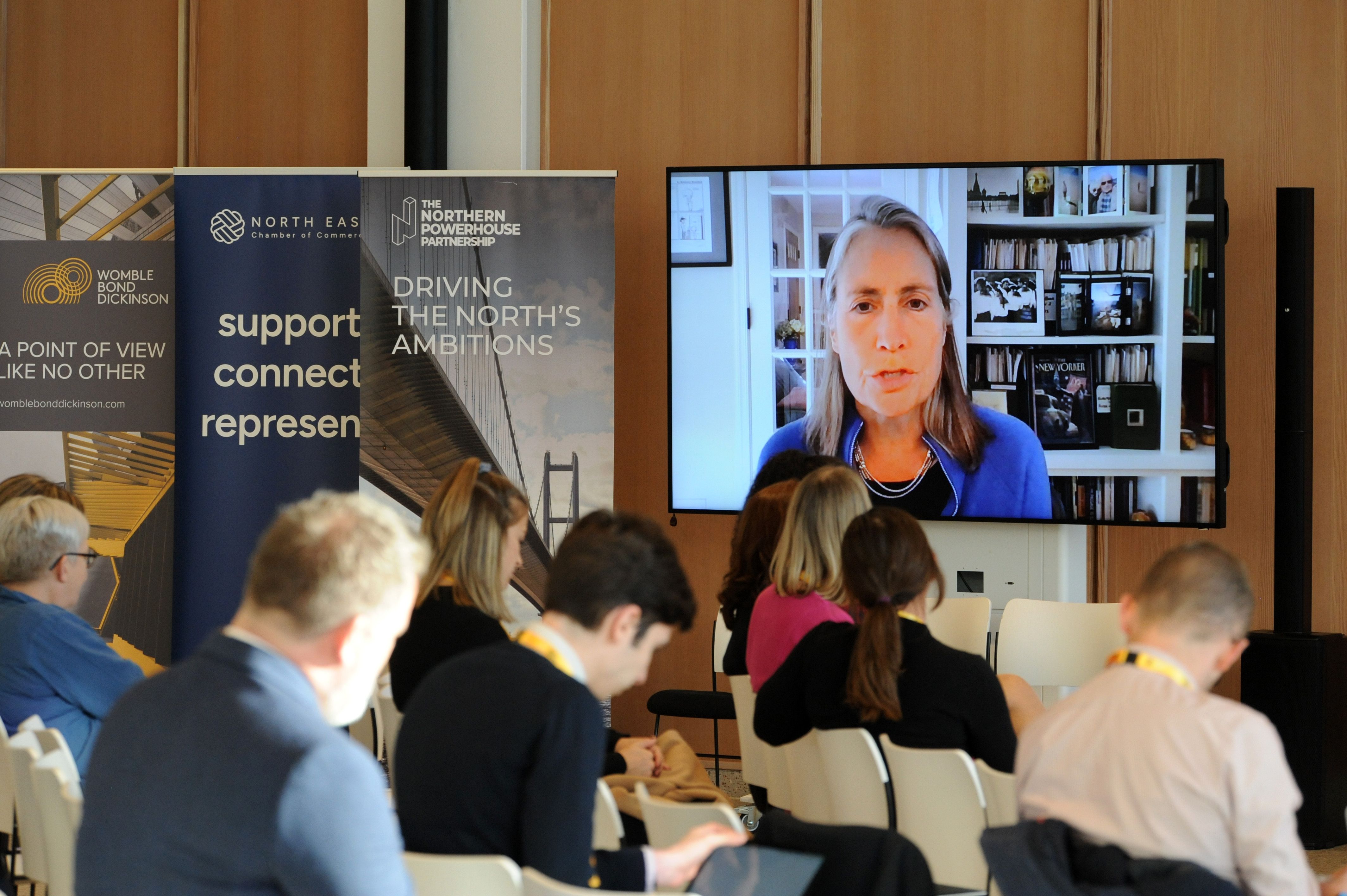 Woman on screen speaking, audience seated in front