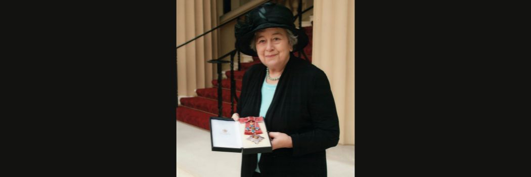 Professor Dame Rosemary Cramp in black hat and jacket holds her Damehood outside Buckingham Palace