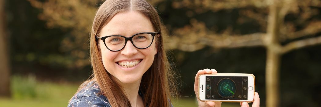 Dr Kelly Jakubowski smiling and holding her mobile phone screen towards the camera against a woodland background