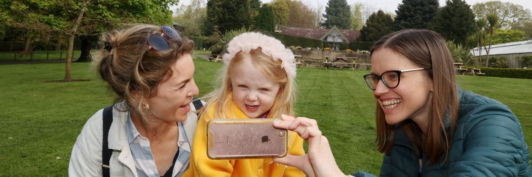 A smiling woman and child look at a mobile phone held by Dr Kelly Jakubowski with a garden view behind them.