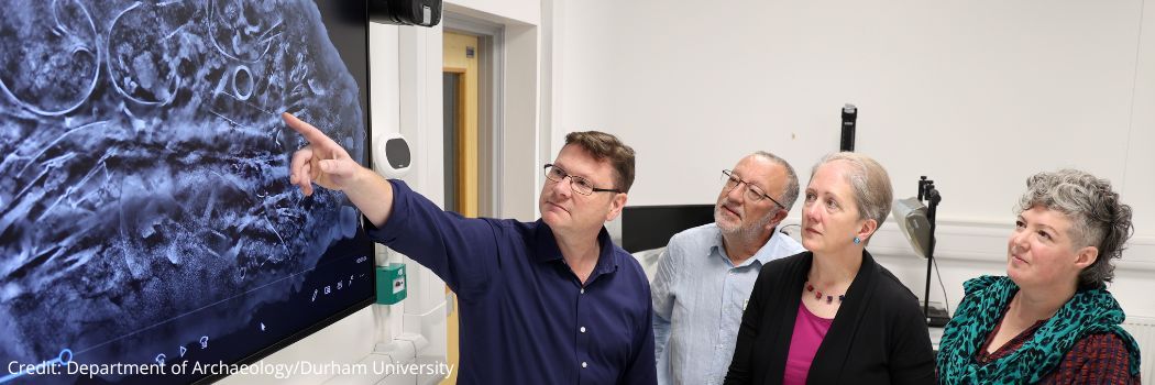 A group of people pointing at CT scan of metal objects on a large display screen.