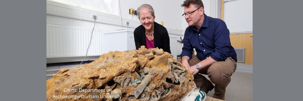 Two people crouching down and looking at a block of tangled metal and soil in a laboratory.