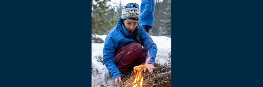 Rosemary Coogan in blue winter coat and woolly hat lights a fire in a snowy woodland