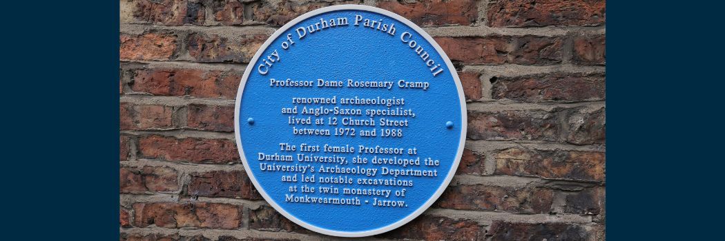 A circular blue metal plaque fixed to a red brick wall. The plaque honours Professor Dame Rosemary Cramp.