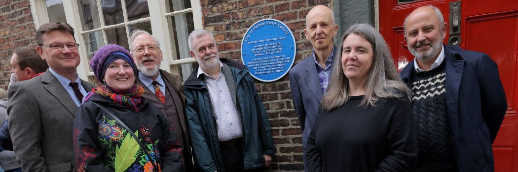 A group of men and women stand in front of a circular blue metal plaque set against a red brick wall.