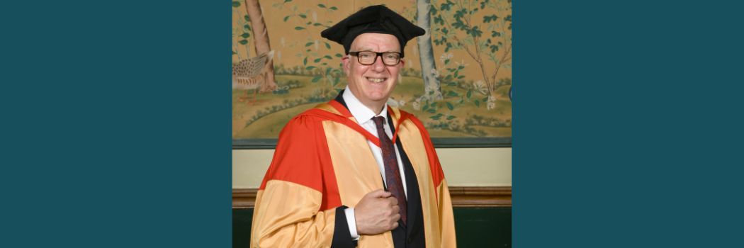 Richard Ovenden wearing a black academic cap and red and gold robes smiling at the camera