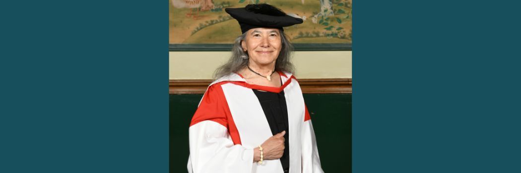 Dalee Sambo Dorough wearing a black academic cap and red and white robes smiling at the camera.