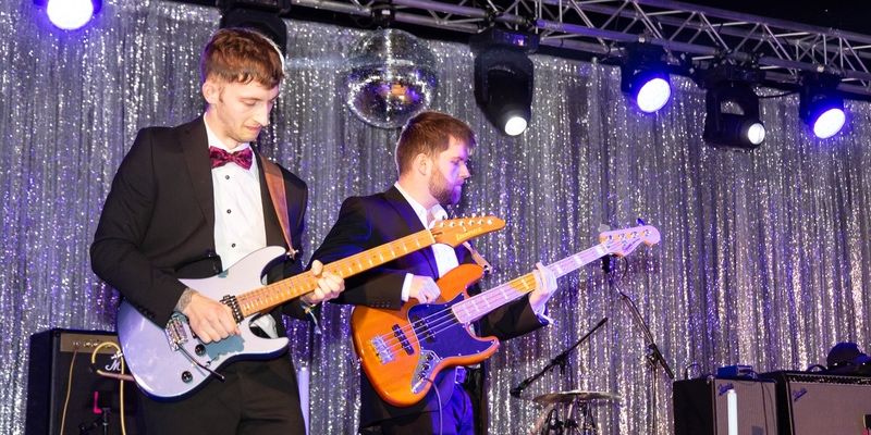 Two students on a stage dressed in black tie playing guitars