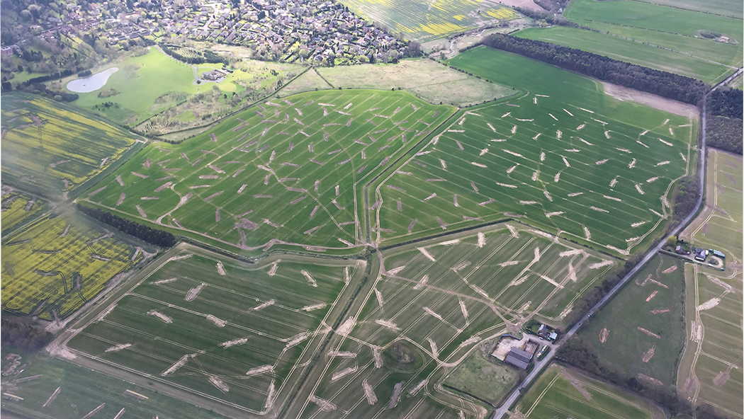 A series of trenches outlined in light soil against the dark green of fields