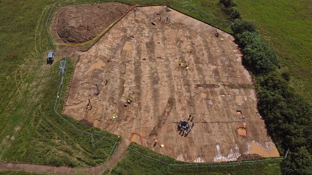 An open air excavation seen from above with features outlined in brown soil and a group of people in a circle as part of an open day