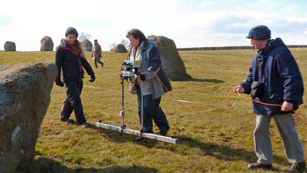 People walking through a field with standing stones carrying geophysics equipment