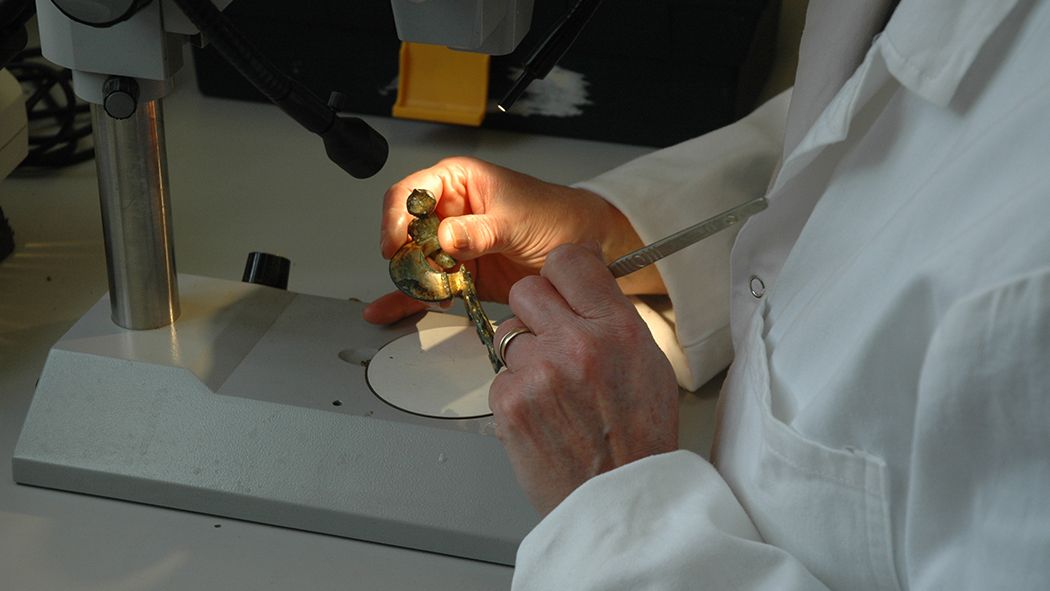 a close-up of an archaeological scientist's hands working on the conservation of a golden brooch