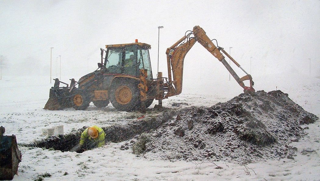 A person in driving snow, crouched in a trench recording a feature with a JCB in the background