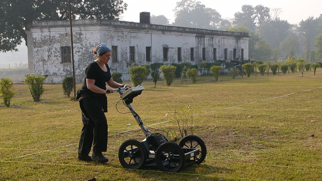 An archaeologist pushing a Ground Penetrating Radar cart with a white building in the background