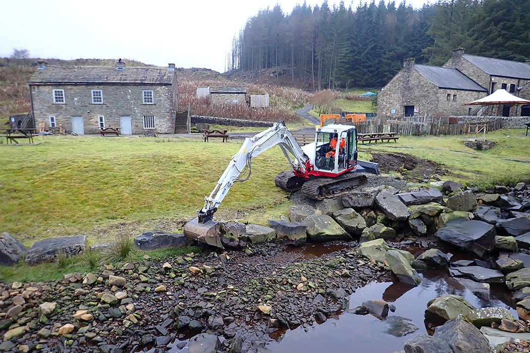 A JCB moving large stone blocks on the edge of a stream, with stone built cottages and forest in the background
