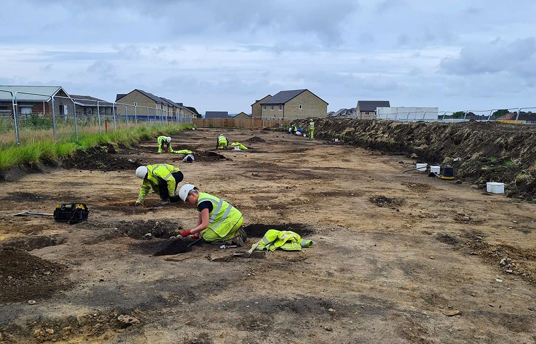 Archaeologists excavating a series of prehistoric pits and ditches with a new housing estate under construction in the background