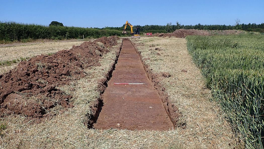 An excavated trench with ranging rod for scale and a JCB in the background