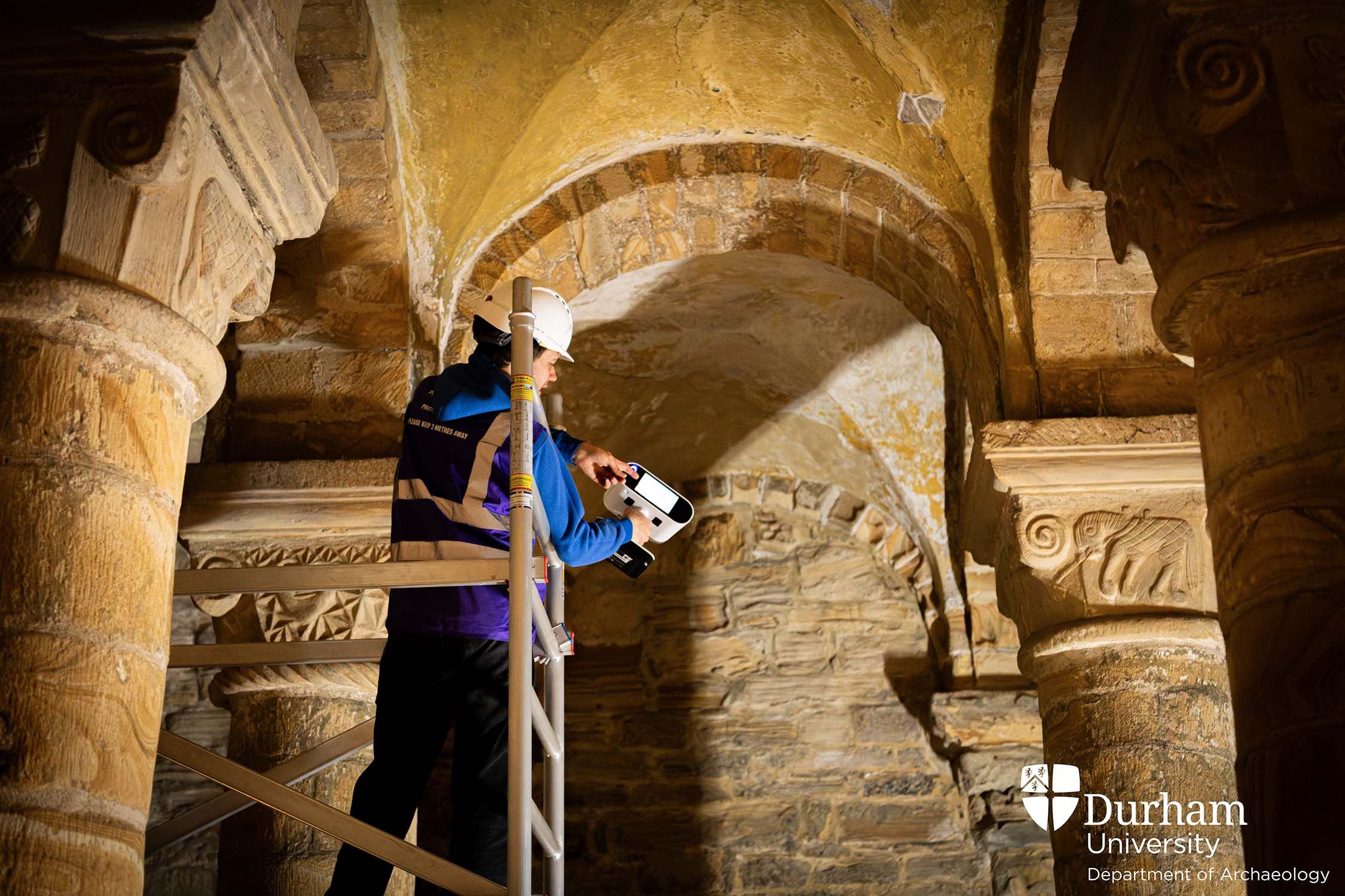 Alexander Jansen standing on a raised platform inside the Norman Chapel, wearing a hard hat and holding a laser scanner.