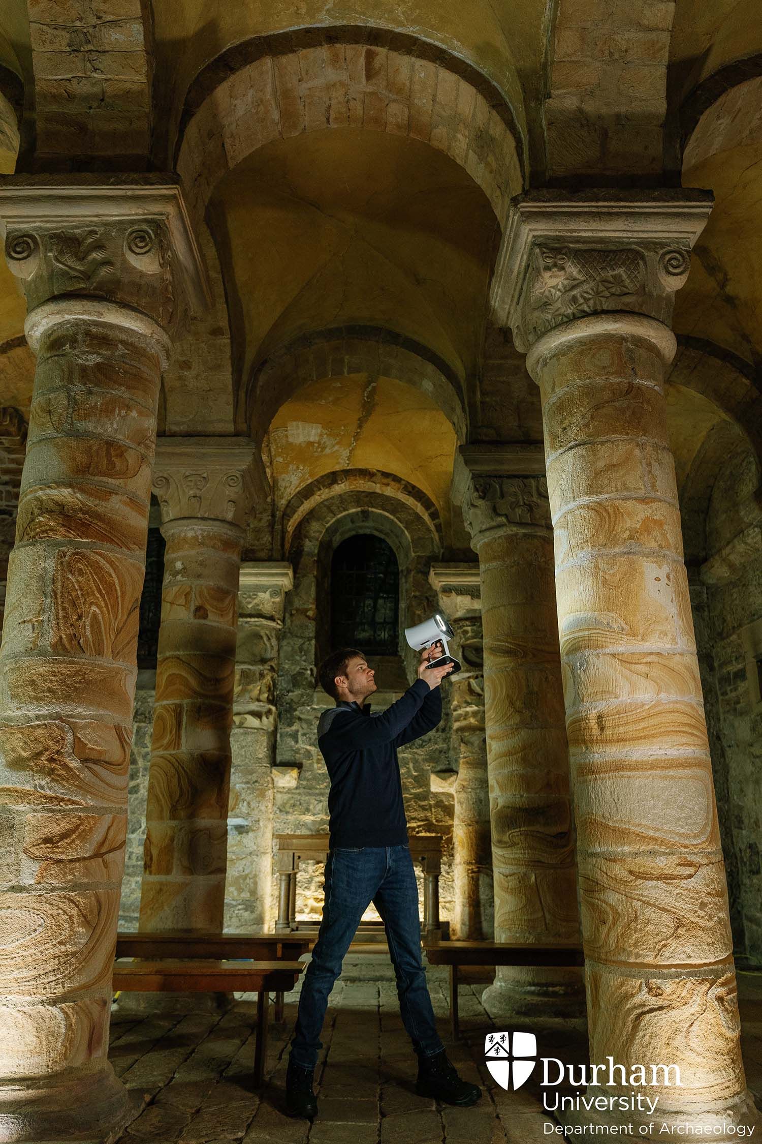 Alexander Jansen standing between four columns inside the Norman Chapel, holding up a laser scanner.