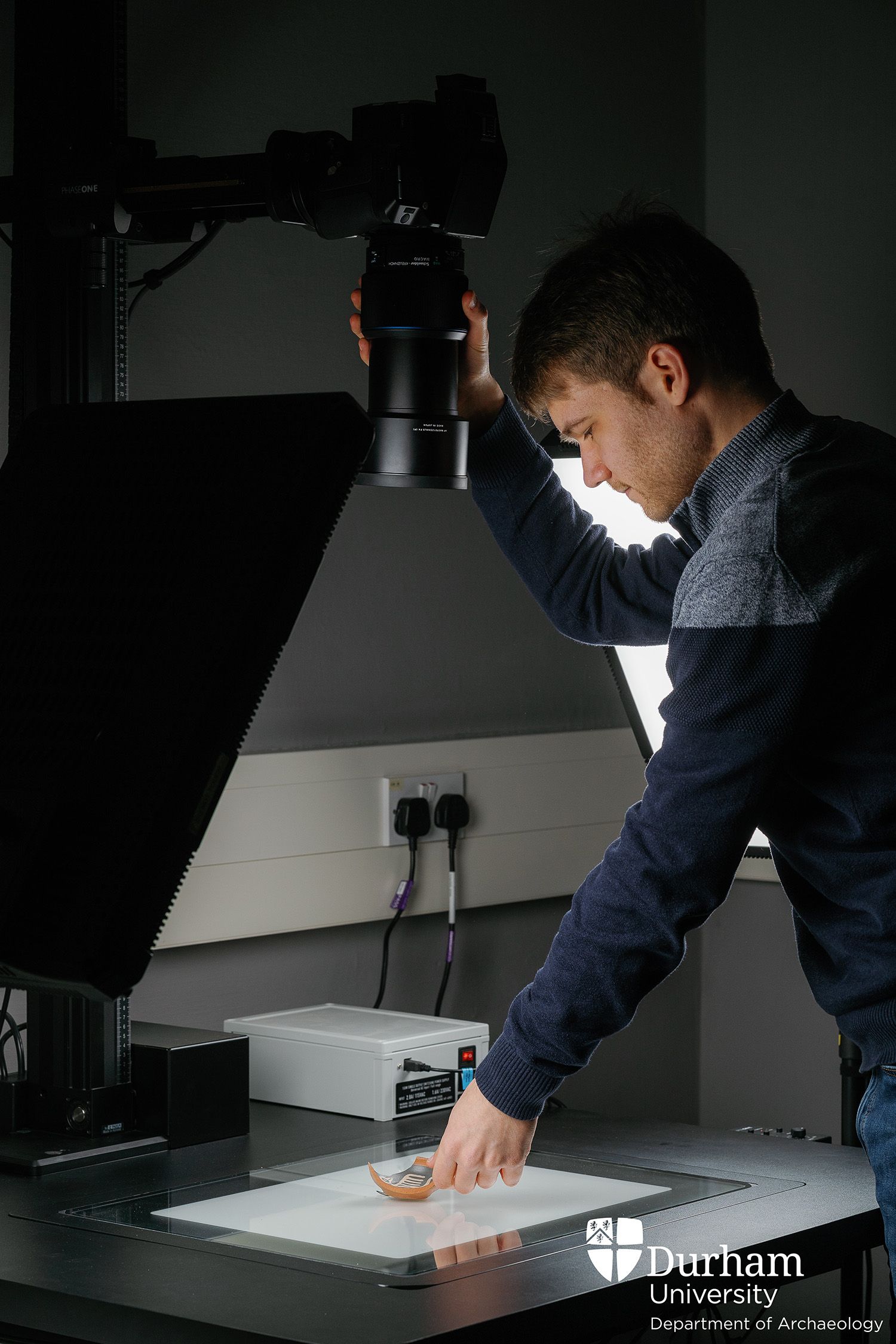 Alexander Jansen stands in a darkened Digital Visualisation Laboratory, using a large camera over an archaeological artefact on the table. There are large lamps on either side of the work surface.