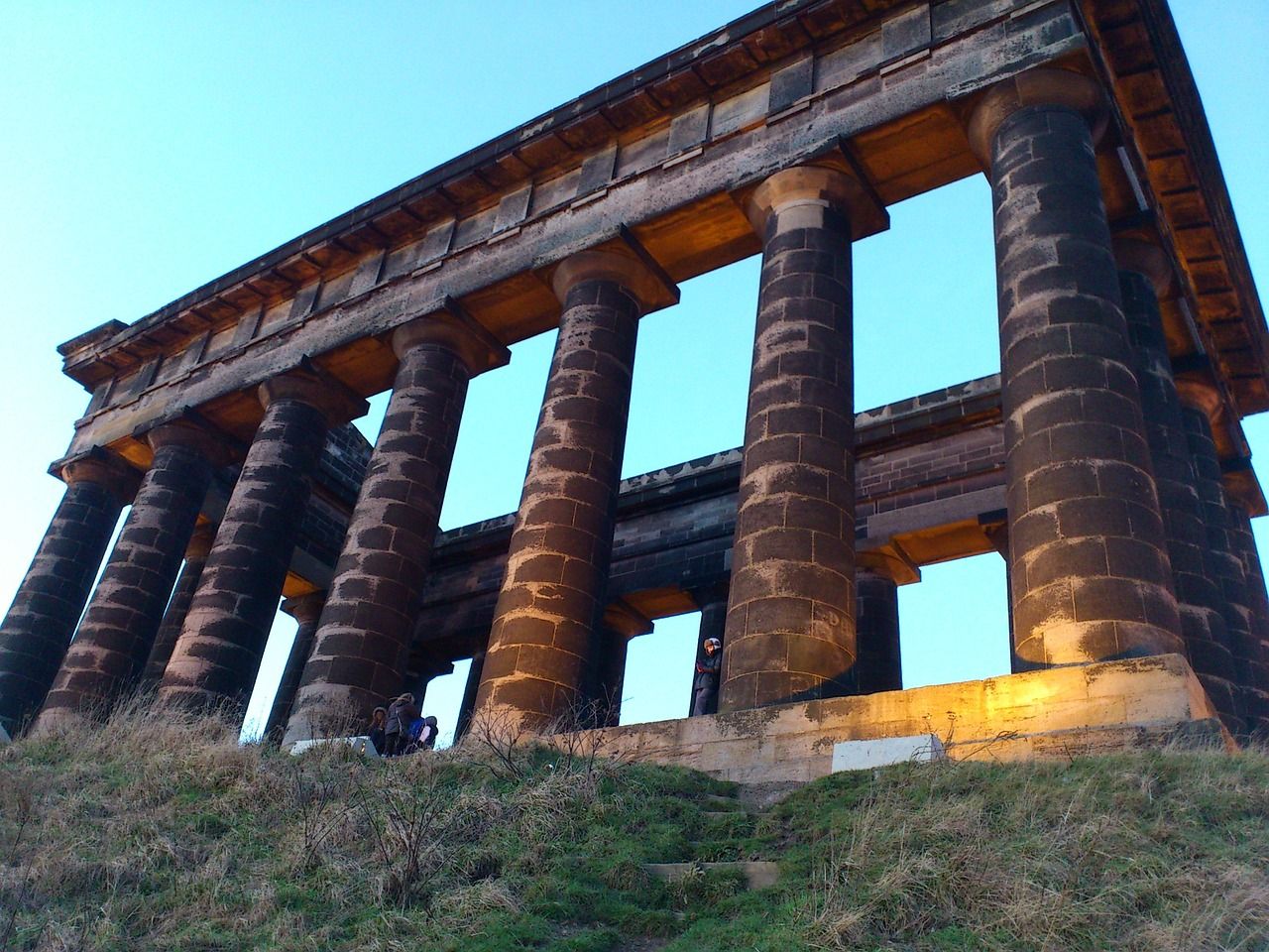 View from below of the side of a replica ancient Greek temple, with seven columns built of dark stone supporting a plain entablature