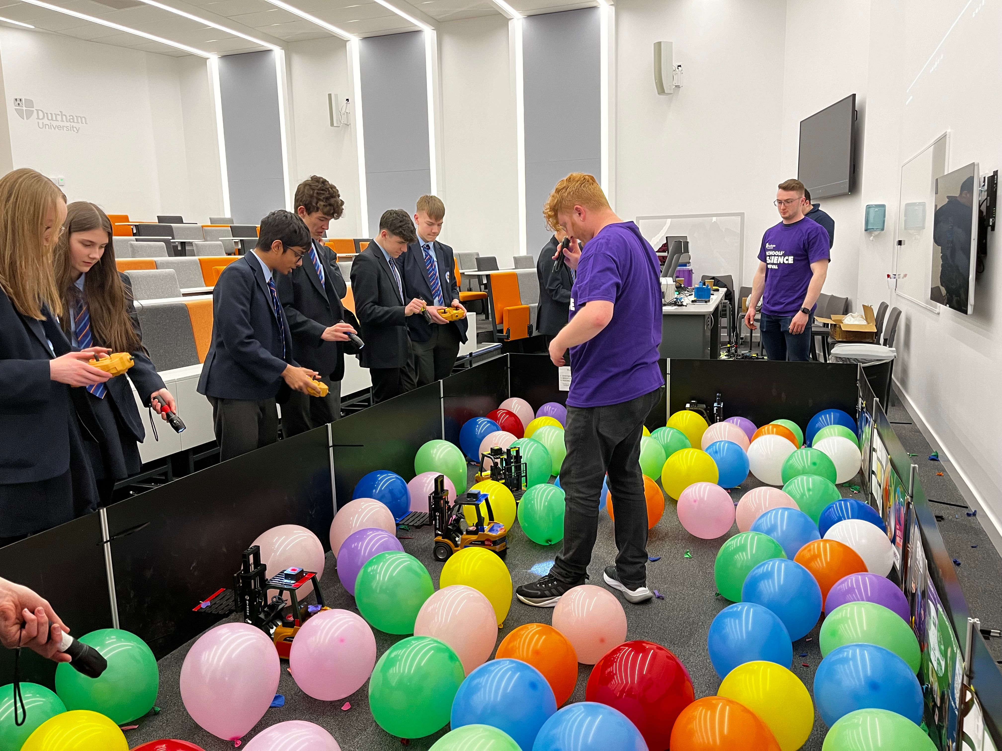 six teenage students in school uniform taking part in science experiment with a large number of multi-coloured balloons on floor
