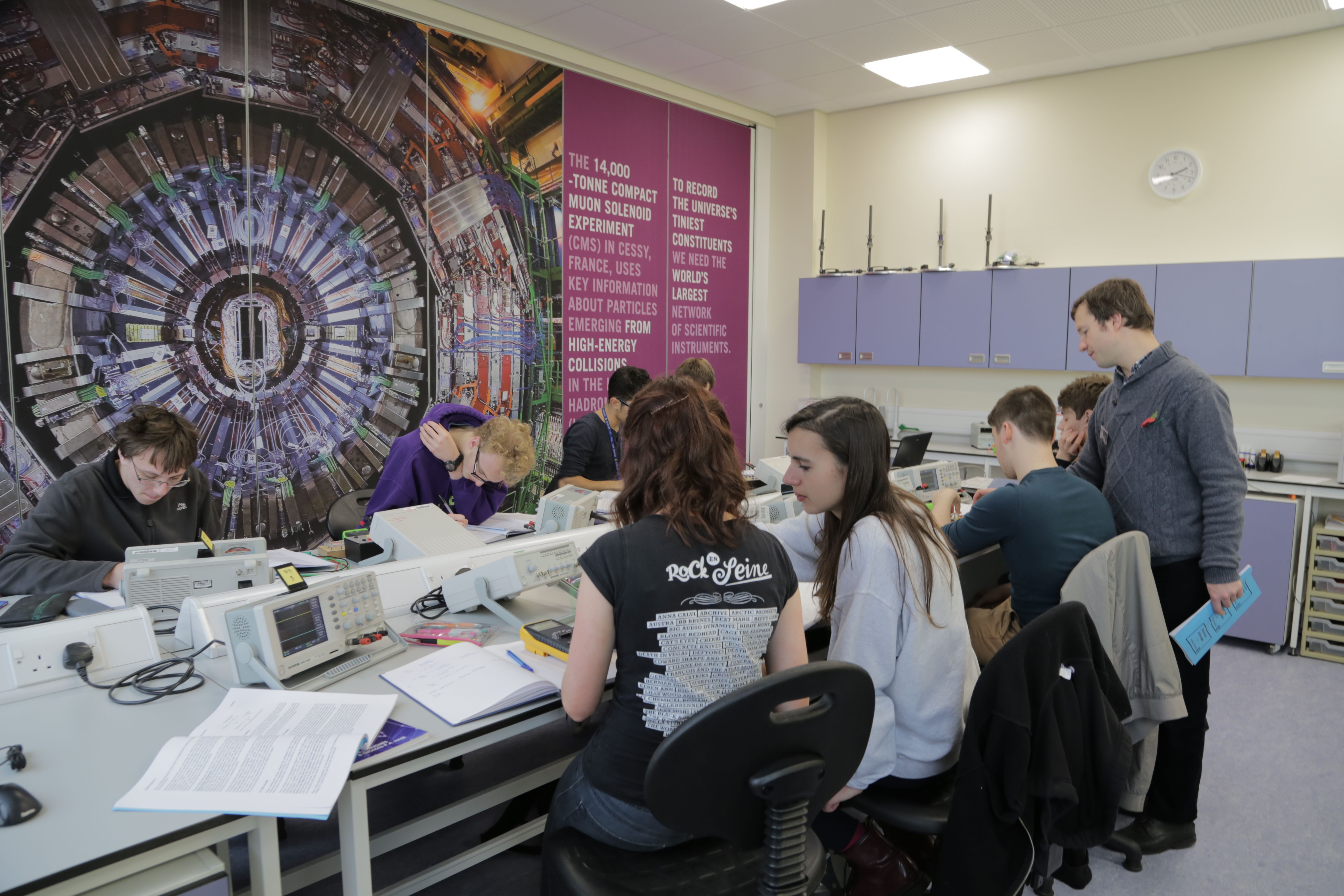 Image of lecturer overseeing group of students at benches in a lab working with oscilloscopes