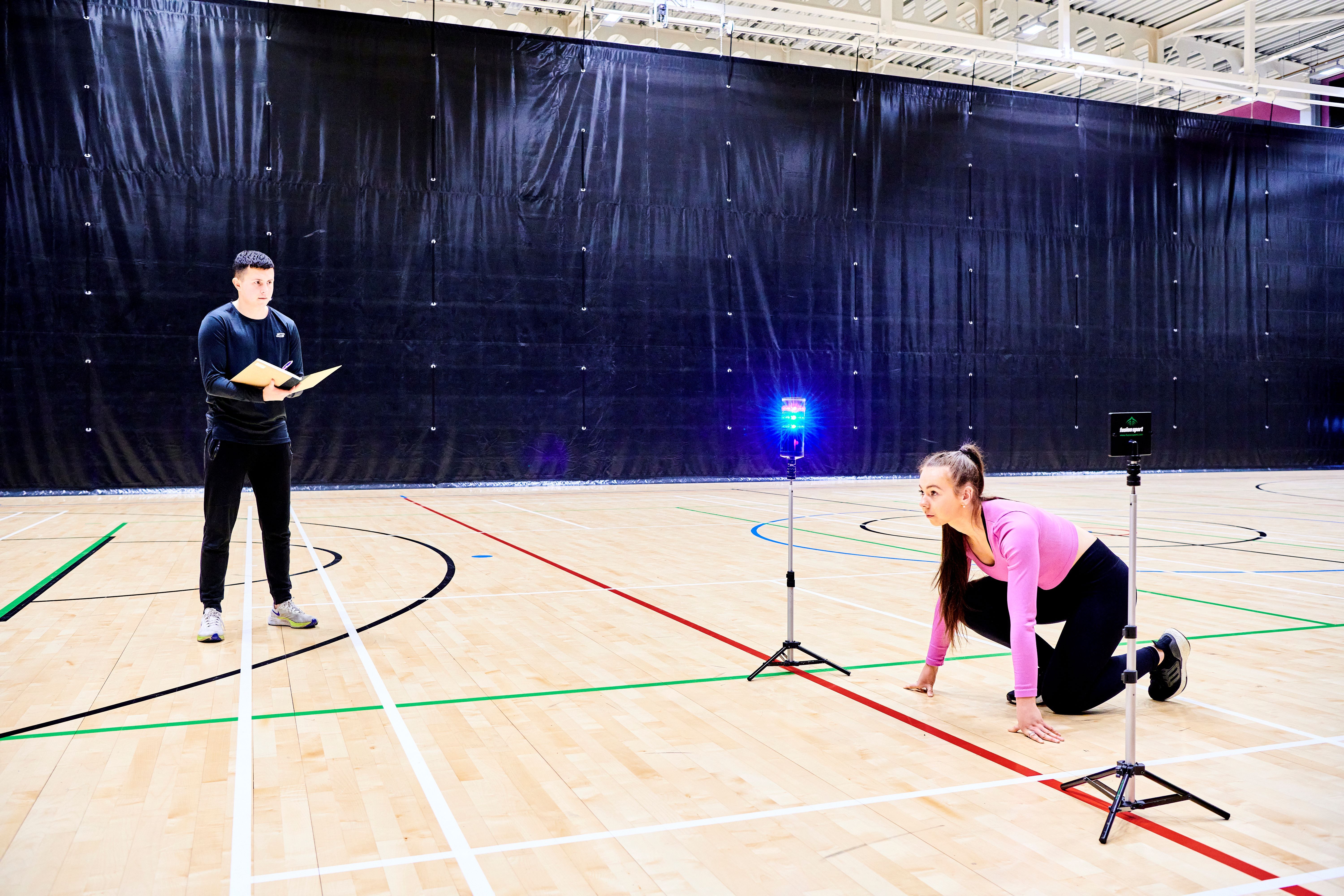 Two students conducting research in Sports Hall. Female student preparing to run while male student records data.