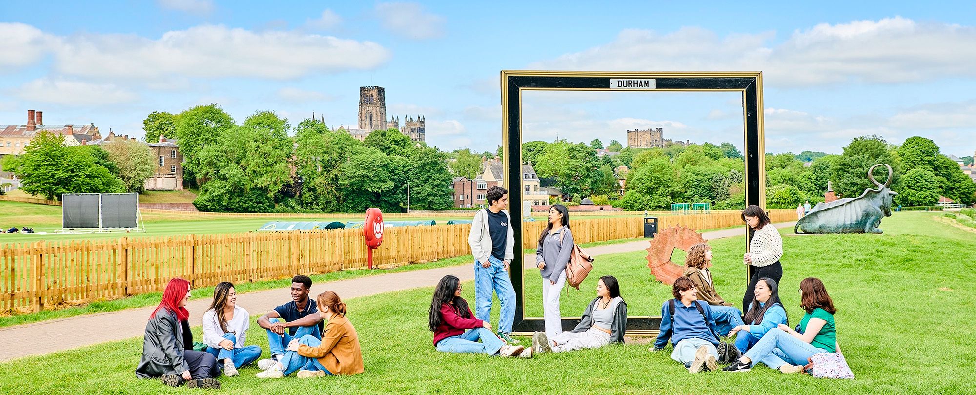 Students sit and stand in groups on the banks of the River Wear. It's a busy shot, with lots going on. They are all dressed in jeans and light jackets. Some are leaning against a large black frame, with 