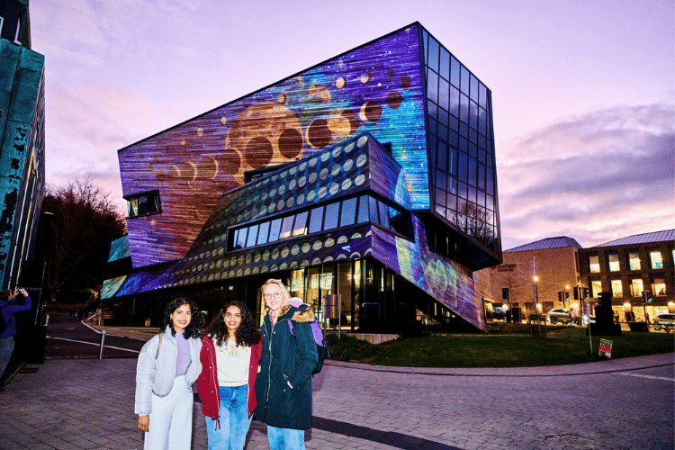 Three students in front of a planetary light art display on the outside of the Ogden Centre