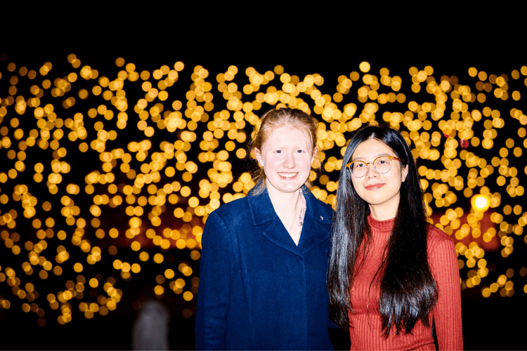 Two female students in front of an illuminated light bulb art display at night