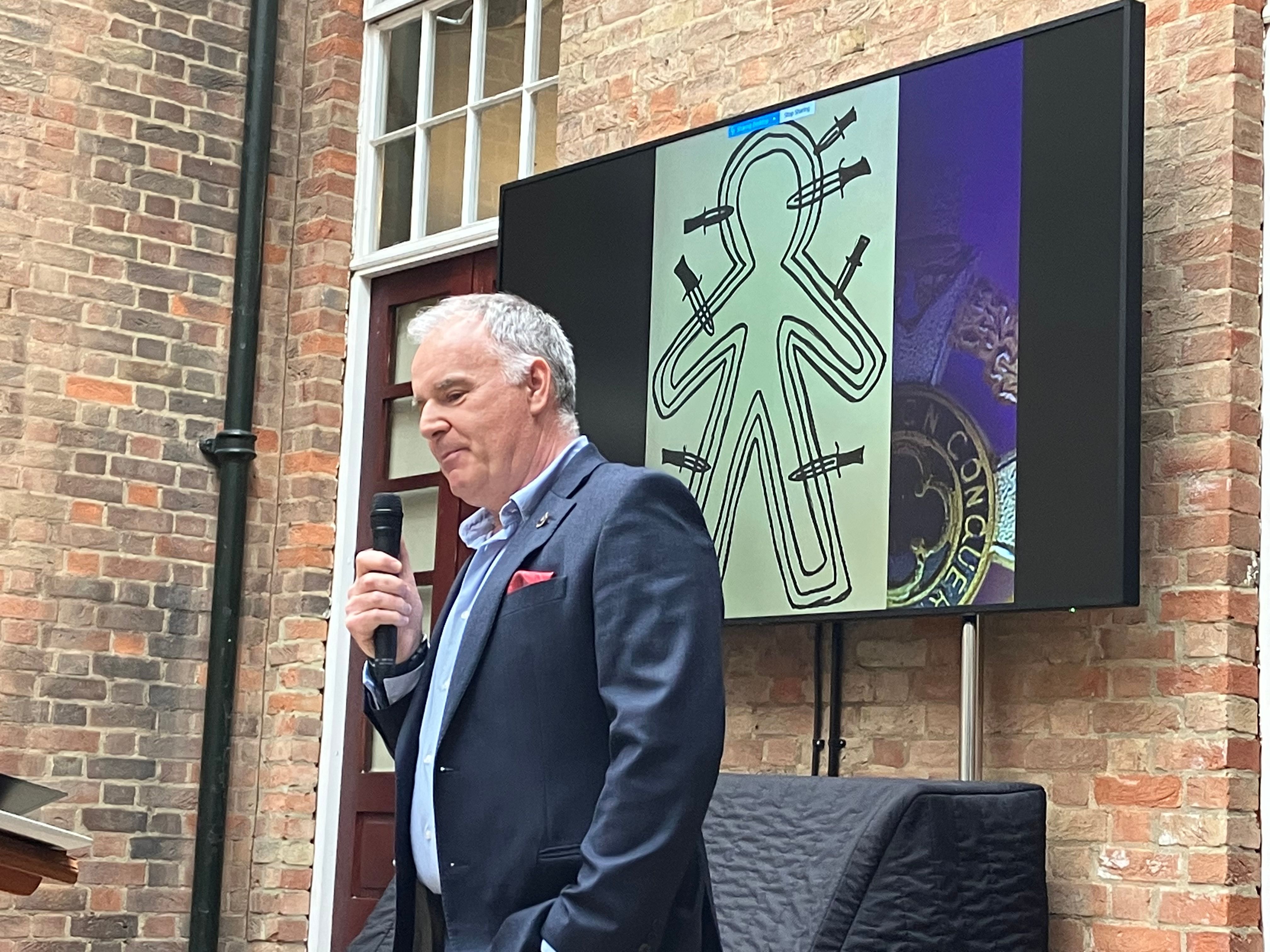 A man speaking in front of a slide showing a line drawing of a person with knives sticking into them