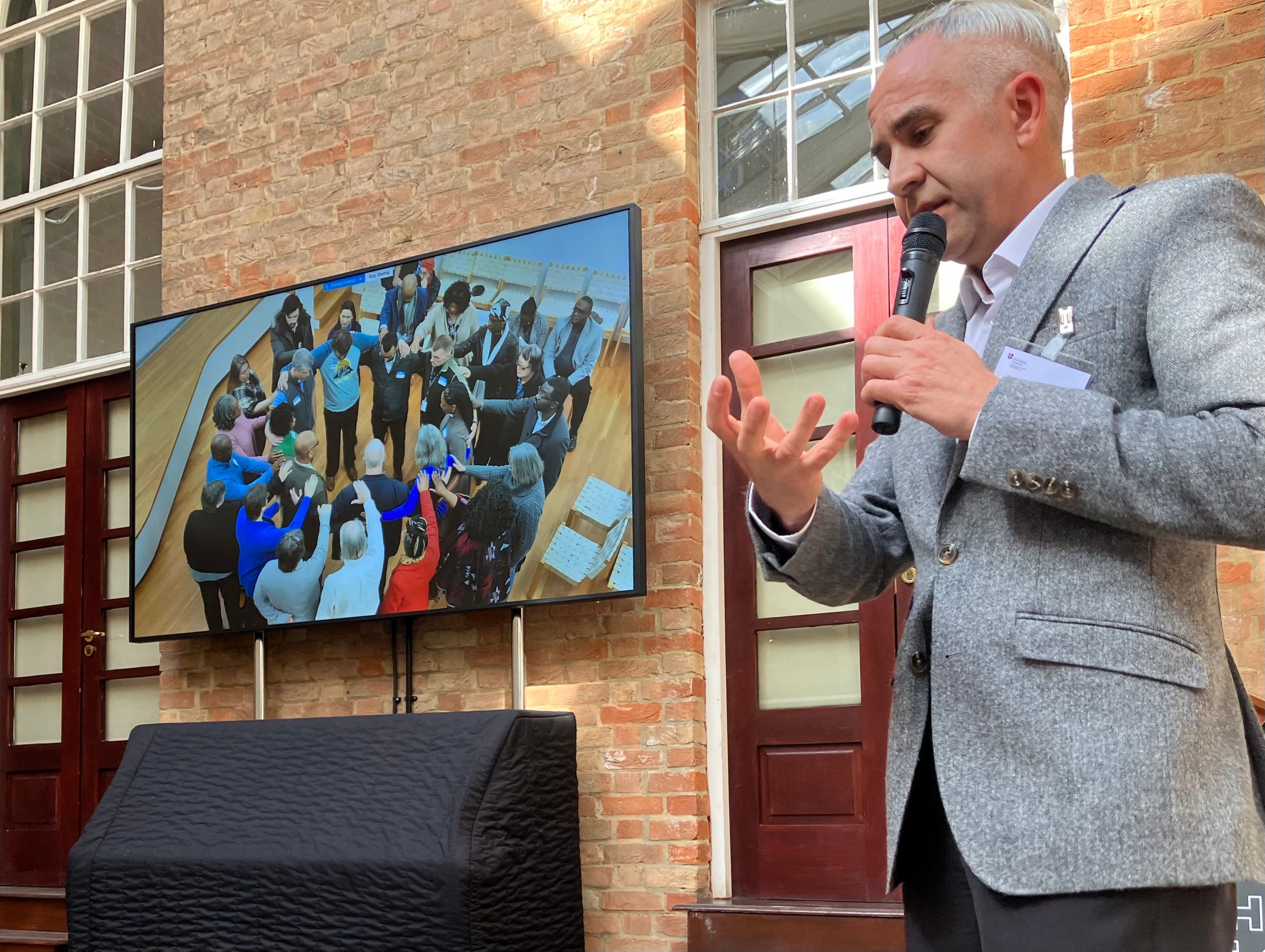 A man talking in front of a slide showing a photo of people gathered round in concentric circles