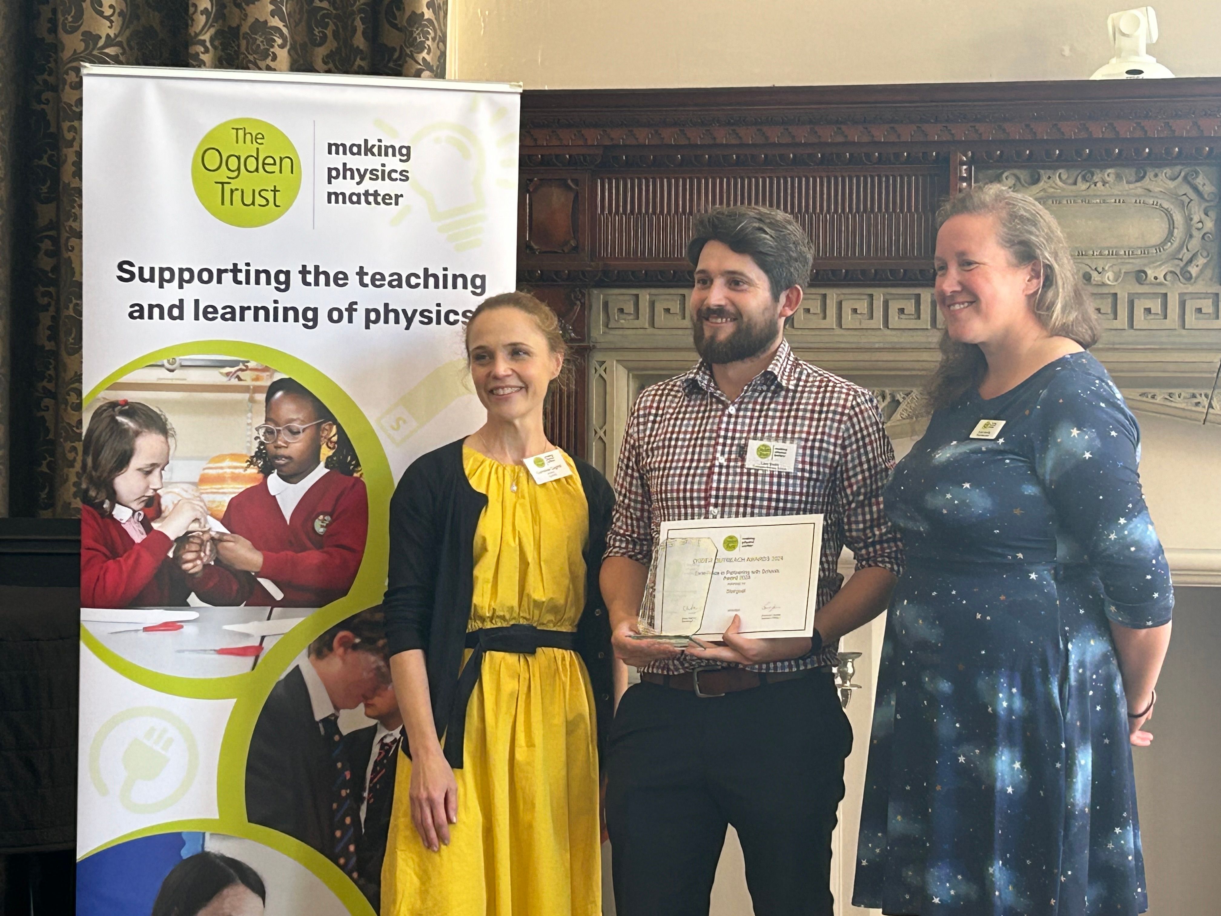 female in yellow dress and male with beard in checked shirt collecting an award made from glass from a second female