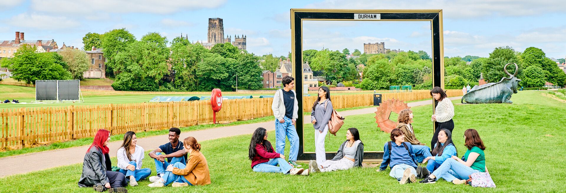 Students sit and stand in groups on the banks of the River Wear. It's a busy shot, with lots going on. They are all dressed in jeans and light jackets. Some are leaning against a large black frame, with 