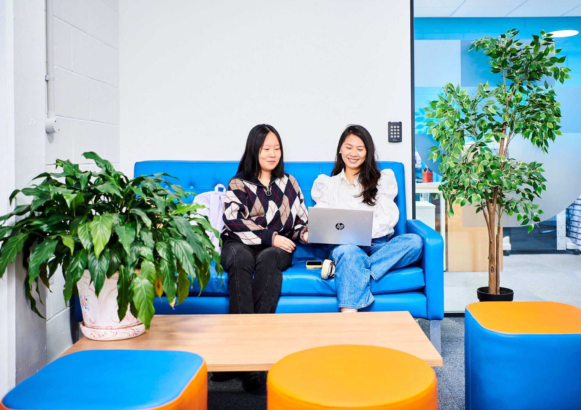 Two female students sit in a reception area. It is brightly lit, with a vivid blue wall, orange and blue furniture, green plants and looks inviting.