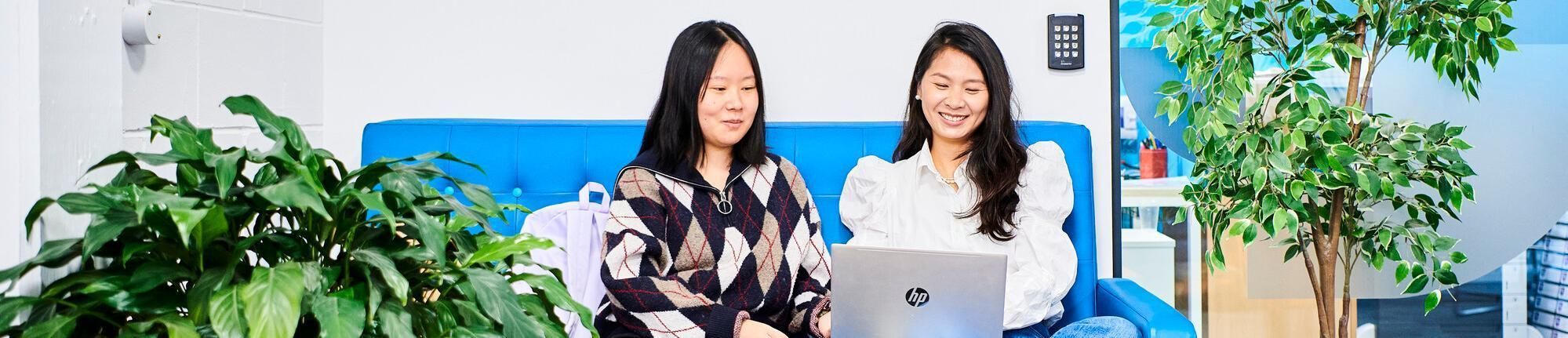 Two female students sit in a reception area. It is brightly lit, with a vivid blue wall, orange and blue furniture, green plants and looks inviting.