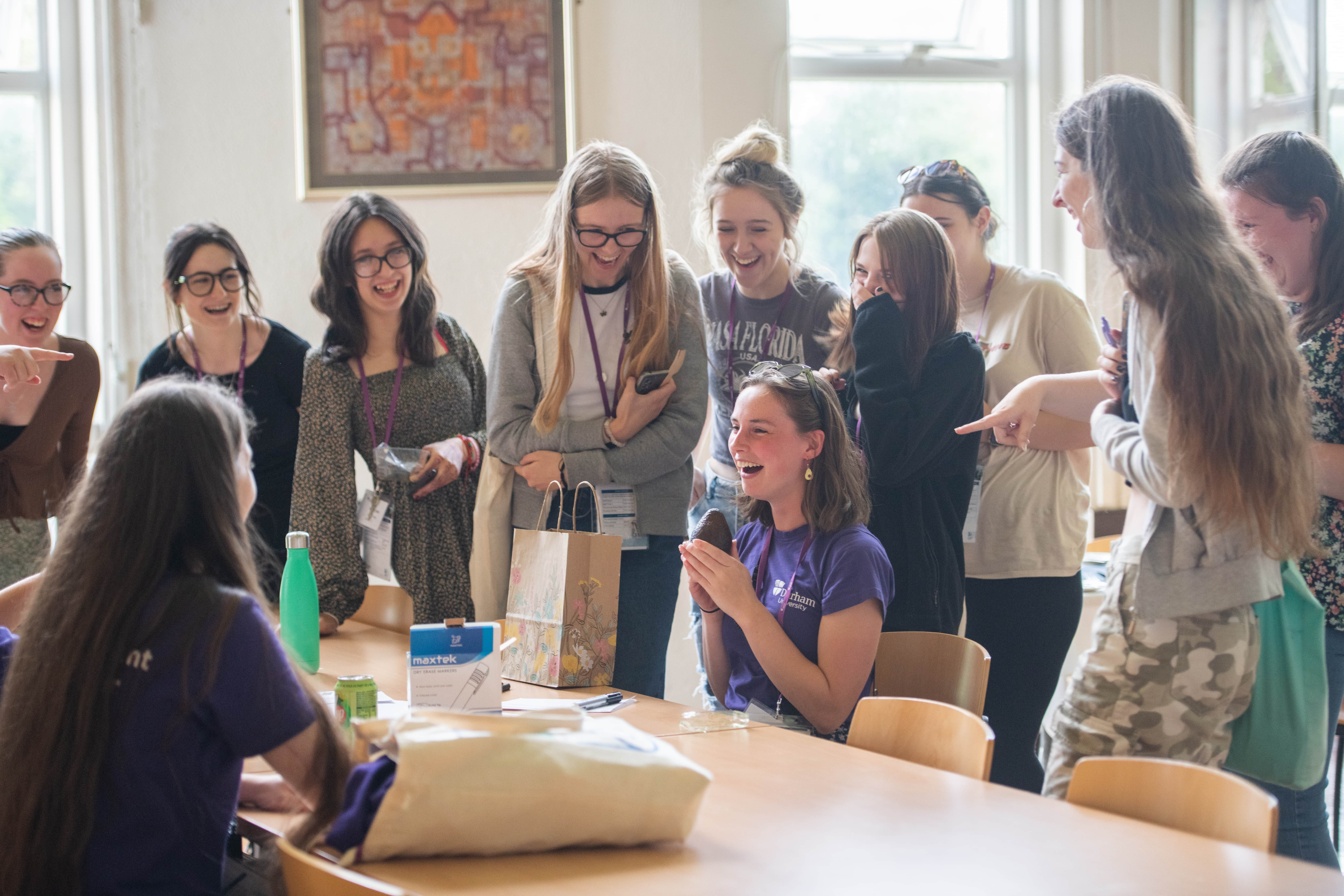 A group of laughing and smiling students huddled round a student ambassador wearing avocado earrings holding a gift of an avocado